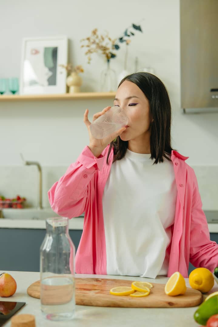 Woman in a pink shirt drinking a glass of water in a bright kitchen